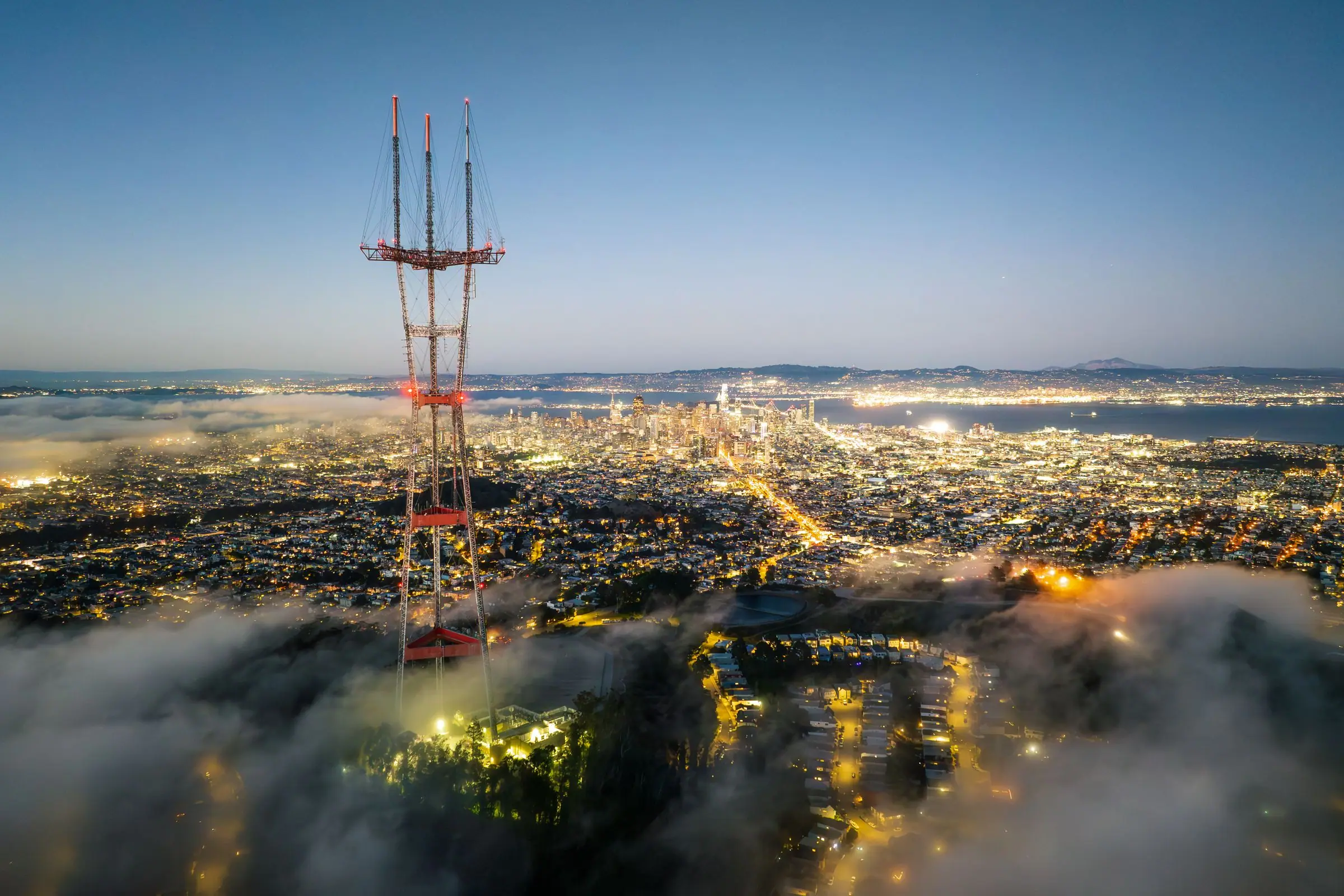Sutro Tower lit above a layer of fog at twilight, with city lights stretching toward downtown
