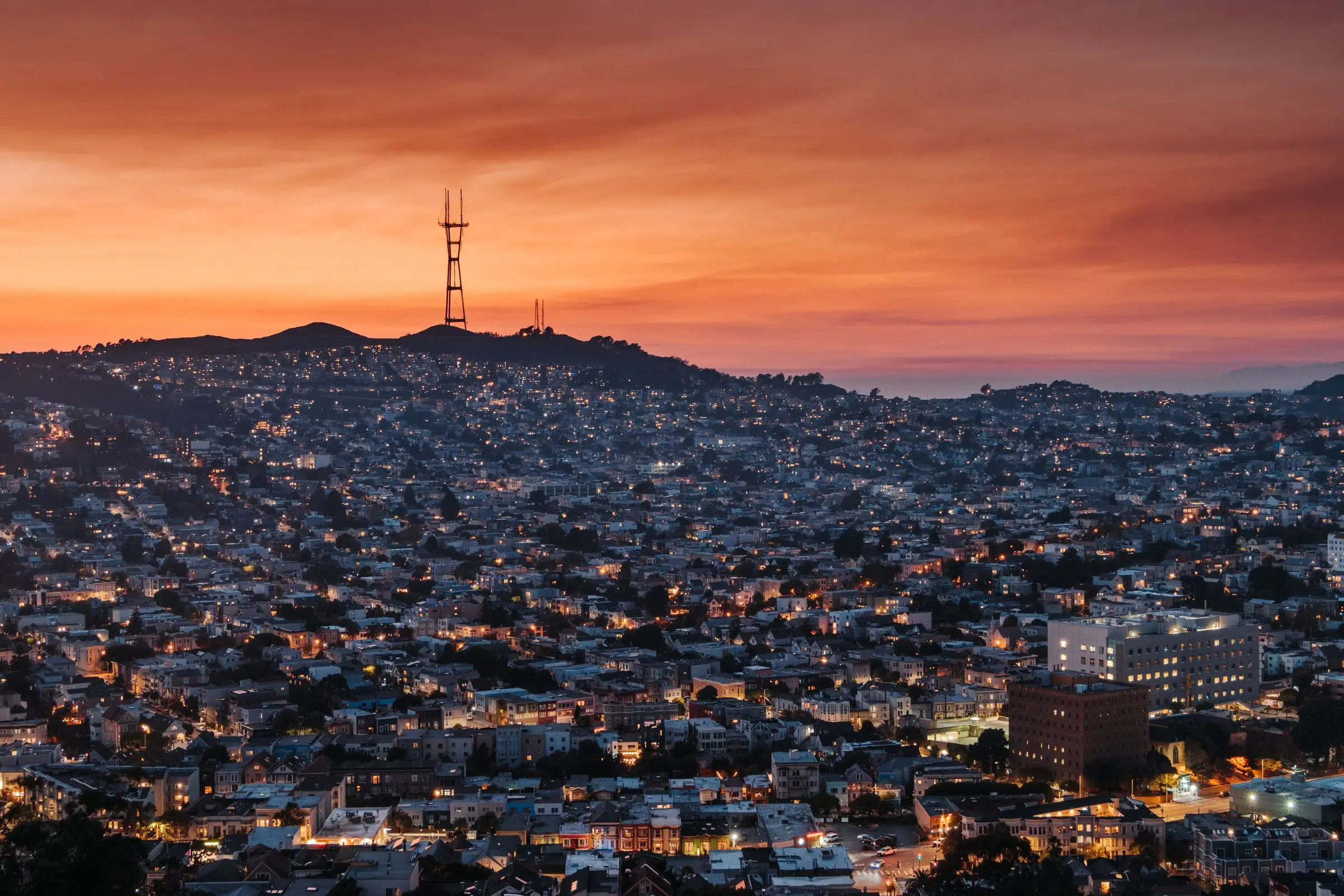 Sutro Tower silhouetted against an orange sunset sky above hillside neighborhoods, with house lights coming on