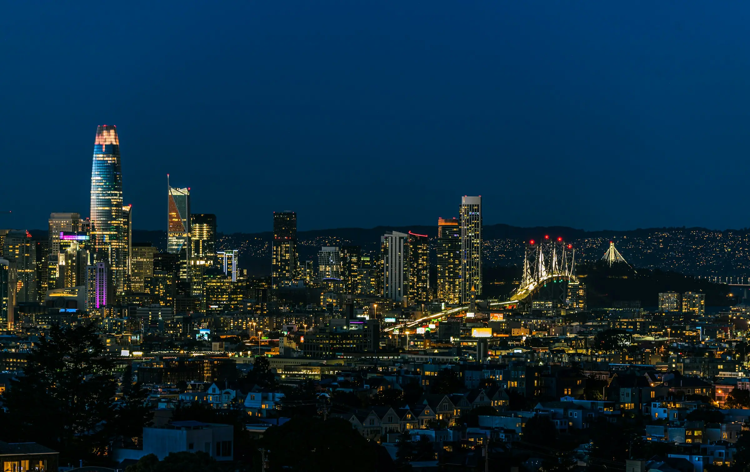 The downtown skyline lit up at blue hour, seen across darkened hillside neighborhoods