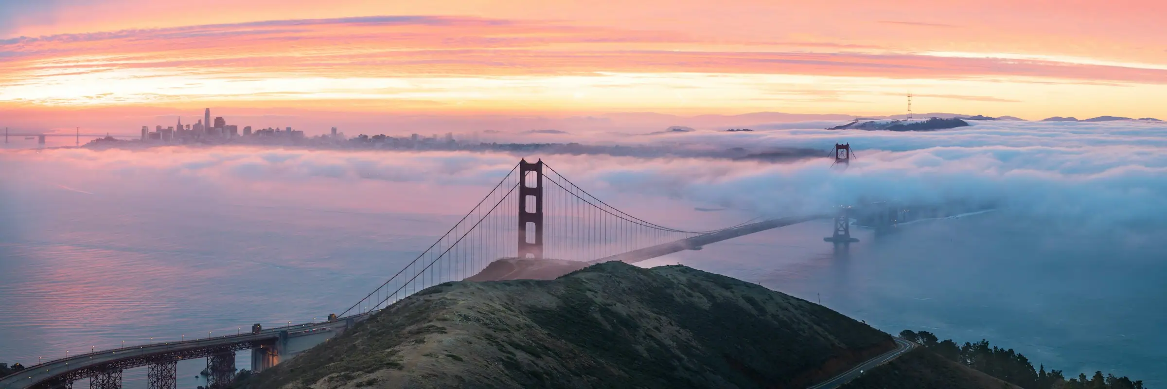 Panorama of the Golden Gate Bridge rising out of a sea of fog at sunrise, with the city skyline on the horizon